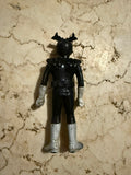 Rear view of the figure placed on a beige marbled surface. The figure wears a black suit with white gloves and boots, and a helmet with red horn-like protrusions. The belt and shoulder sculpting are visible, emphasizing the figure’s tokusatsu design lineage.