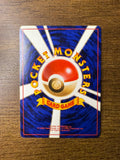 Back side of a Japanese Pokémon trading card placed on a wooden surface, showing a central Poké Ball with radiating white beams on a blue background. Yellow curved text reads “POCKET MONSTERS,” with a yellow ribbon labeled “CARD GAME.” Includes 1996 copyright information.