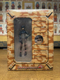 Front view of the Full Metal Panic! DVD limited edition box with a desert camouflage design, showing a detailed mecha illustration through a clear plastic window, photographed on a wooden surface with illustrated figures in the background.