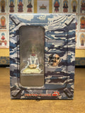 Japanese Full Metal Panic anime camouflage-patterned DVD box on wooden table in front of Buddhist shrine with statues. Clear front window shows small seated figure of green-haired boy in school uniform on glowing base. Text reads "フルメタル・パニック!" and DVD logo.
​