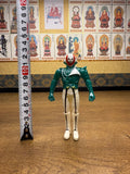 Naval Commander Bosgun sofubi figure from Kamen Rider Black RX (1988), standing upright next to a vertical measuring tape showing a height of approximately 10 cm. The figure wears a green top, white pants with black vertical stripes, and a red-and-white skull-like mask. The static pose and scale reference emphasize its compact size and collectible nature.