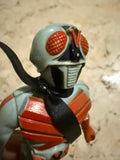 Close-up of the Kamen Rider X figure’s head and upper body, placed on a beige marble surface. The helmet has large red eyes and a vented mouthpiece. The chest features red armor with light blue accents and a black scarf. The sculpting and paintwork reflect vintage tokusatsu styling.