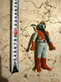 Overhead view of the Kamen Rider X figure lying next to a measuring tape, indicating a height of approximately 13 cm. The figure wears a light blue suit with red boots, gloves, and chest armor, and a black scarf around the neck. The helmet has red eyes and a mouthpiece. Measuring tape includes markings “GS,” “KISAKI,” “JIS B 7512,” and “COMMAX.”