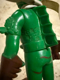 Close-up of the underside of a toy figure’s feet. The left foot is engraved with “アスカ” (Asuka), possibly referencing a character or manufacturer. The right foot has an oval stamp reading “MADE IN CHINA,” suggesting a later or alternate production run. The figure’s green outfit and brown boots are partially visible, though this may not be the original Popy release.