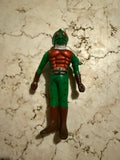 Full-body view of the Kamen Rider Amazon sofubi figure lying on a marbled surface. The figure features a green body, red chest armor, brown gloves and boots, and a helmet with red eyes and antennae. A white scarf wraps around the neck. The sculpt includes muscle definition and a belt, reflecting classic tokusatsu hero design from the Showa era.