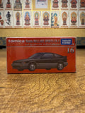 Tomica Premium TP16 Honda Ballade Sports CR-X box featuring red packaging and car illustration, placed against a wall decorated with framed spiritual figures. Includes branding and commemorative Japanese text.