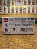 Bottom side of the Tomica Premium product box showing Japanese and English product details, barcode, safety certifications, and Takara Tomy contact info. Background features a wall of labeled Buddhist figure illustrations.
