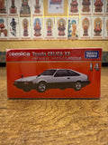 Front of the Tomica Premium No. 14 Toyota Celica XX box featuring a silver model car illustration and branding. Background includes a golden wall with grid-arranged Buddhist figure cards, adding cultural depth to the scene.