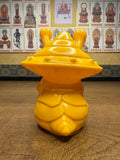 Yellow plastic Ultra-Luck Money Box Kanegon figurine resembling a stylized snail or alien with a segmented body and two antennae-like eye stalks. A coin slot is visible on its back, indicating its function as a money box. The figure is placed on a wooden surface, with a background wall featuring framed Buddhist icons labeled in Chinese characters, including a central red-robed figure surrounded by deities."