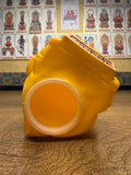 Bottom view of the yellow plastic Ultra-Luck Money Box Kanegon figurine lying on its side on a wooden surface. The base features a circular opening, likely for inserting or removing coins. Embossed text reads 'CHINA' and 'ITEM NO. MADE IN CHINA.' In the background, a wall displays framed images of Buddhist deities with Chinese or Japanese labels, including a central figure in red robes with a golden border."