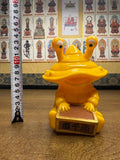 Yellow plastic figurine of Ultra-Luck Money Box Kanegon with three protruding eyes, seated on a wooden surface and holding a rectangular box with Japanese characters '吉千両' meaning 'Good fortune and a thousand ryō.' A vertical measuring tape beside the figure shows a height of approximately 17 cm. The background includes framed images of Buddhist figures labeled in Japanese, enhancing the cultural contrast.
