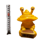Yellow plastic figurine of Ultra-Luck Money Box Kanegon, a cartoonish frog-like kaiju with two large eyes on stalks and a wide mouth. The character is holding a rectangular brown chest with Chinese characters '青千両' printed on the front, symbolizing 'Green Thousand Ryo'—a reference to wealth. The figurine stands on a wooden surface next to a vertical measuring tape, indicating a height of approximately 10 cm. The design is whimsical and culturally themed, blending playful aesthetics with prosperity symbolism