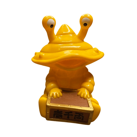 Yellow figurine of Ultra-Luck Money Box Kanegon, featuring exaggerated eye stalks, a wide mouth, and a cartoonish body. The character holds a rectangular box with a wood-textured surface and Chinese characters '青千両' on the front, referencing traditional currency and prosperity. The figure is made of plastic and placed on a wooden surface, showcasing a playful yet culturally inspired design.