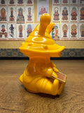 "Yellow figurine of Ultra-Luck Money Box Kanegon designed with a spiral shell-like head, large eyes, and dressed in a robe. The character is seated and holding a book, suggesting a scholarly or meditative pose. The background features a wall of framed Buddhist deities labeled in Chinese text, blending playful design with spiritual iconography."