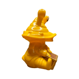 Seated yellow figurine of Ultra-Luck Money Box Kanegon with a large head, single protruding eye stalk, and wide open mouth. The character holds a small rectangular object resembling a chest or book with a brown surface and gold edges. The stylized, cartoonish design and exaggerated features reflect Kanegon’s playful kaiju identity, making it a visually striking collectible.