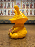 Yellow plastic figurine of Ultra-Luck Money Box Kanegon seated on a wooden surface, featuring two eye stalks, a wide mouth, and exaggerated cartoonish features. The character holds a brown rectangular chest or book. Behind it, a wall displays golden-hued framed images of Buddhist figures labeled in Chinese script, creating a contrast between the whimsical toy and traditional religious artwork."