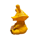 Yellow plastic figurine of Ultra-Luck Money Box Kanegon in a seated position, featuring a large conical head with two eye stalks protruding from the top. The character holds a brown rectangular chest with both hands. The design combines snail-like and alien elements, with a smooth, whimsical finish that emphasizes Kanegon’s quirky personality and money-themed motif.