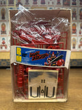Open box of the Red Thunder plastic model kit showing red sprue parts, a sticker sheet, and packaging insert labeled “1:100 Scale All Plastic Model Kit.” Background includes a wall of collectible figurine images.