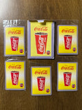 "Set of Five Coca-Cola-themed playing cards arranged on a wooden surface. Each card features a red Coca-Cola cup filled with ice and soda against a yellow background, with 'Enjoy Coca-Cola' written at the top. The border has a blue and white checkered pattern, and a circular 'Drink Coca-Cola' logo appears in the bottom right. The central card includes a flap, suggesting it is part of the packaging."