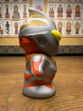 Side profile of the Ultraman Gaia piggy bank figure in a confident, heroic stance with arms bent and hands on hips. The figure has a chibi-style design with a large head and small body, painted in silver with red and gold accents. Bright yellow eyes and a sculpted helmet crest add character detail. The exaggerated proportions and pose reflect a playful reinterpretation of the iconic Ultraman hero.
