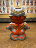 Close-up front view of the Ultraman Gaia piggy bank, showcasing a futuristic helmet with a pointed crest and smooth, featureless face. The figure is painted in orange, gold, and silver tones, with the embossed text “Daiwa Bank” prominently displayed on the chest. This indicates the item may have been a promotional tie-in or branded collectible, merging corporate sponsorship with pop culture appeal.