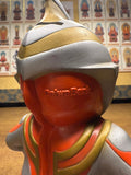 Back view of a stylized Ultraman Gaia piggy bank designed in a Daruma doll-inspired form. The figure features a rounded body with silver, orange, and gold coloring, and a helmeted head with a crest. Embossed text “Daruma” is visible on the back, blending traditional Japanese design with tokusatsu aesthetics. The figure’s cartoonish proportions and smooth finish suggest it’s a soft vinyl or molded plastic collectible from 1998.