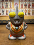 Frontal view of the Ultraman Gaia piggy bank in a standing pose with hands on hips. The figure features a silver body with red and gold highlights, large yellow eyes, and a blue triangular emblem on the chest. The exaggerated head and simplified body proportions reflect a chibi-style design. The confident stance and vibrant paintwork emphasize its appeal as a playful yet iconic tokusatsu collectible.