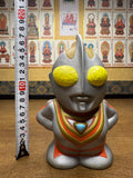 Front-facing view of the Ultraman Gaia piggy bank placed beside a vertical measuring tape, indicating a height of approximately 10 centimeters. The figure has large yellow eyes, a silver helmet with crest, and a red-and-gold suit with a blue triangular emblem on the chest. The stylized proportions and bright colors highlight its chibi aesthetic, while the measurement confirms its compact collectible size.