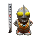 Frontal view of the Ultraman Gaia piggy bank in a standing pose with hands on hips. The figure features a silver body with red and gold highlights, large yellow eyes, and a blue triangular emblem on the chest. The exaggerated head and simplified body proportions reflect a chibi-style design. The confident stance and vibrant paintwork emphasize its appeal as a playful yet iconic tokusatsu collectible.