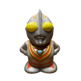 Frontal view of the Ultraman Gaia piggy bank in a standing pose with hands on hips. The figure features a silver body with red and gold highlights, large yellow eyes, and a blue triangular emblem on the chest. The exaggerated head and simplified body proportions reflect a chibi-style design. The confident stance and vibrant paintwork emphasize its appeal as a playful yet iconic tokusatsu collectible.