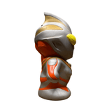 Side profile of the Ultraman Gaia piggy bank figure in a confident, heroic stance with arms bent and hands on hips. The figure has a chibi-style design with a large head and small body, painted in silver with red and gold accents. Bright yellow eyes and a sculpted helmet crest add character detail. The exaggerated proportions and pose reflect a playful reinterpretation of the iconic Ultraman hero.