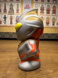 Side view of the Ultraman Gaia piggy bank, featuring a metallic silver body with red and gold accents and a large yellow eye. The figure has a cartoonish, oversized head and compact body. Embossed text “Coin Bank” is visible on the side, confirming its dual function as a savings container and collectible figure. The design blends utility with pop culture styling.