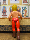 Rear view of the Golden Mask figure standing on a wooden floor, facing a wall decorated with framed Buddhist deity illustrations. The figure has yellow hair, red trunks, and brown boots. The juxtaposition of the vintage toy with traditional religious artwork creates a striking contrast between pop culture and spiritual iconography.