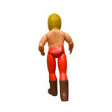 Rear view of the Golden Mask sofubi figure standing on a flat surface. The figure has molded yellow hair, a shirtless muscular torso, red trunks over red pants, and brown boots. The arms are slightly bent, and the figure is posed in a neutral stance. The plastic shows minor wear, consistent with vintage age.