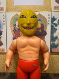 Frontal view of the Golden Mask figure with exaggerated yellow mask and red pants, placed against a background of Japanese religious artwork. The mask features wide eyes and sharp teeth. The backdrop includes labeled illustrations of Buddhist deities, enhancing the cultural depth of the image.