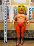 Upright view of the Golden Mask figure standing beside a measuring tape, showing a height of approximately 30 cm. The figure has a yellow mask, red pants, and brown boots. The background features multiple illustrated panels of Buddhist deities with Japanese text, creating a visual contrast between vintage toy design and traditional spiritual art.