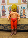 Upright view of the Golden Mask figure with shirtless torso, red pants, brown boots, and a fierce yellow mask. The figure is placed against a backdrop of framed Buddhist deity illustrations with Japanese labels. The toy shows signs of wear, including scuff marks on the legs and torso, consistent with vintage condition.