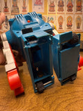 "Close-up of a vintage blue toy robot with red and silver components, showing an open front compartment that reveals internal wiring and a small motor. The robot includes wheels, gears, a red button on top, and vent-like molded details. It is placed on a wooden surface with a background wall featuring a grid of illustrated cultural or religious figures in traditional attire, each labeled in East Asian script."