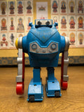 "Vintage toy robot standing on a wooden surface, primarily blue with red and silver arms and a boxy retro design. The robot features visible screws, mechanical detailing, and a head with two protruding circular elements resembling eyes and a ridged panel suggesting a mouth. Its rectangular legs have small wheels, indicating mobility. In the background, a wall is covered with framed images of Buddhist figures or deities, each labeled with text in Tibetan or Sanskrit script. The scene contrasts the mechanical