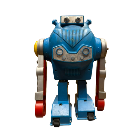 "Vintage toy robot with a blue boxy body, silver and red jointed arms, and black wheels at the base. The head features two protruding antennae and a face panel with circular and rectangular shapes resembling eyes and a mouth. The design evokes mid-century fascination with space-age robotics and mechanical toys."