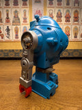 "Vintage toy robot standing on a wooden surface, primarily blue with silver and red arms and yellow circular accents on the sides of its head. The robot has a boxy body with rivet-like details, a face featuring two round eyes and a grill-like mouth, and small black wheels on its feet. Behind the robot is a wall covered with framed images of Buddhist figures arranged in a grid, each labeled with text in East Asian script, creating a contrast between mechanical design and spiritual iconography."