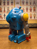 "Vintage toy robot with a rounded blue body, yellow and silver arms, and red claw-like hands, standing on a wooden surface. The robot features mechanical details such as molded vents and bolts, evoking a retro sci-fi aesthetic. In the background, a wall displays framed illustrations of seated Buddhist figures, each accompanied by Chinese text, highlighting a juxtaposition between pop culture and traditional spirituality."