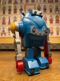 "Vintage toy robot with a rounded blue body and a single mechanical arm ending in a red claw-like gripper. The arm is silver and red, and the body includes molded mechanical details such as vents. The robot stands on a wooden surface, with a background wall covered in framed religious or spiritual illustrations labeled in Japanese or Chinese characters, creating a visual contrast between technology and tradition."