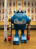 "Vintage blue toy robot standing upright on a wooden surface next to a vertical measuring tape showing a height of approximately 20 cm. The robot has a boxy torso, cylindrical silver and red arms, and rectangular legs with wheels. Its face includes two circular eyes and a metallic panel with bolts, enhancing its mechanical appearance. The background features a wall of individually framed religious or spiritual figures, likely Buddhist, creating a visual juxtaposition between nostalgic technology and sacred 