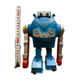"Vintage toy robot standing upright next to a vertical measuring tape indicating a height of approximately 20 cm. The robot has a blue body, red feet, silver arms, and a face panel with circular 'eyes' and button-like features. The design includes wheels on the feet and retro mechanical detailing."