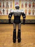 Complete rear view of the Shadowmoon sofubi figure from Kamen Rider Black (1988), featuring a metallic silver and black color scheme. The figure stands upright with clenched fists, wearing a helmet with dual antennae and sculpted armor plating across the shoulders, arms, and legs. The design reflects the biomechanical aesthetic of the character, emphasizing its villainous presence and collectible appeal.
