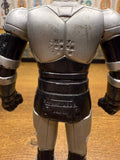 Rear view of a headless Shadowmoon sofubi figure from Kamen Rider Black (1988), showcasing a muscular silver and black sculpt with detailed armor plating on the arms, torso, and legs. Embossed Japanese text on the upper back reads “© 石森プロ ・東映動画 東 映,” and the lower back is marked “BANDAI 1987 JAPAN,” confirming its official licensing and production origin. The absence of the head highlights the figure’s modular design or wear, relevant for condition grading and authenticity verification.