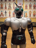 Side profile of the Shadowmoon sofubi figure from Kamen Rider Black (1988), posed in a combat-ready stance with clenched fists. The figure features segmented silver armor with black highlights, green eyes, and matching green accents on the belt and lower legs. The helmet includes antennae and a sculpted crest, reinforcing the character’s futuristic and insectoid design. Ideal for showcasing the figure’s silhouette and dynamic proportions.