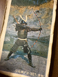 Vintage printed card featuring a character dressed in traditional samurai armor, posed dynamically with legs apart and arms extended while aiming a long bow with orange and black segments. The figure wears a helmet with a crest, chest plate, and full arm and leg protection. A quiver is visible on the back. The background shows rocky terrain and a stream, suggesting an outdoor setting. Japanese copyright text at the bottom reads “©石森プロ・NET 東映,” linking the image to Ishimori Productions, NET, and Toei—indicat