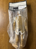 Close-up of the Shapley Alien figure inside a transparent plastic bag with a header card labeled “Outsart.” Japanese text includes “シブレー星人” (Shiburē seijin), translating to “Sibley Alien.” The figure is gold with white accents on the limbs and torso, and wears a silver helmet with a grid-like texture. The packaging suggests a specialty or limited-run item from a Japanese toy line.